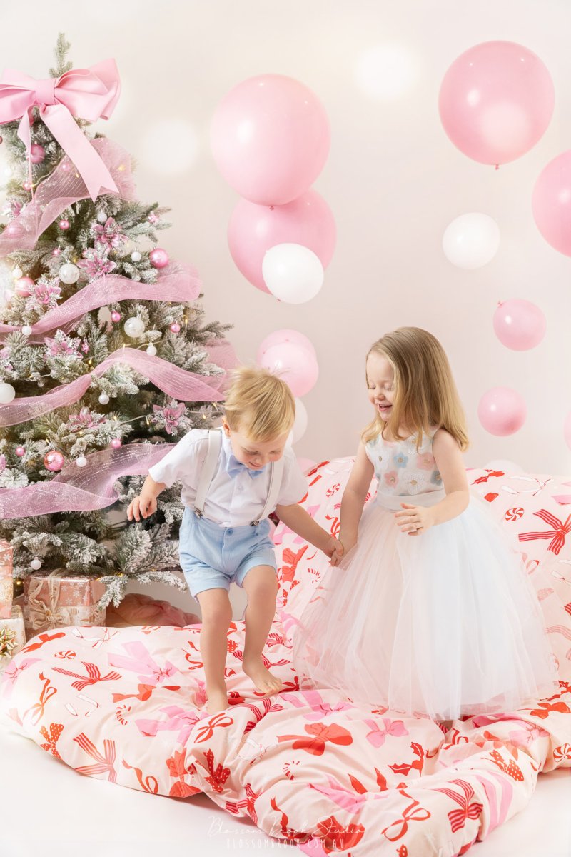 Two children holding hands and playing during a Christmas Family Photoshoot, surrounded by pink decorations and festive styling, captured as Children Christmas Photography in a bright studio.