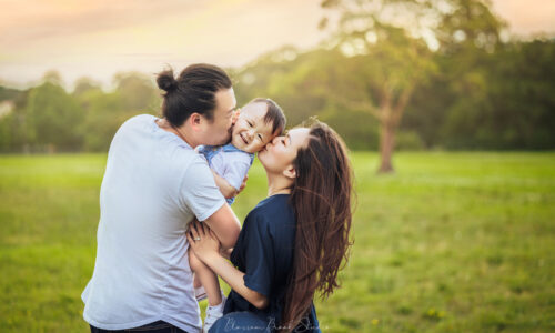 Gallery | Family photoshoot | Blossom Brook Studio 54 Loving parents kissing their smiling child during an outdoor family photoshoot in a beautiful green park at sunset.