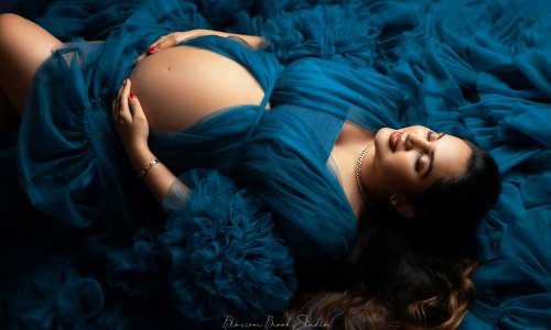 Newborn Photography Blog | Blossom Brook Studio 606 A maternity photoshoot of a couple on a deep blue background. The woman wears a flowing teal gown with ruffles, showcasing her baby bump, while her partner sits beside her, gently holding her belly and kissing her temple. Captured by Blossom Brook Studio.